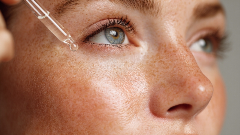 Krupan kadar lica žene sa pegicama dok nanosi serum iz pipete na kožu pored oka