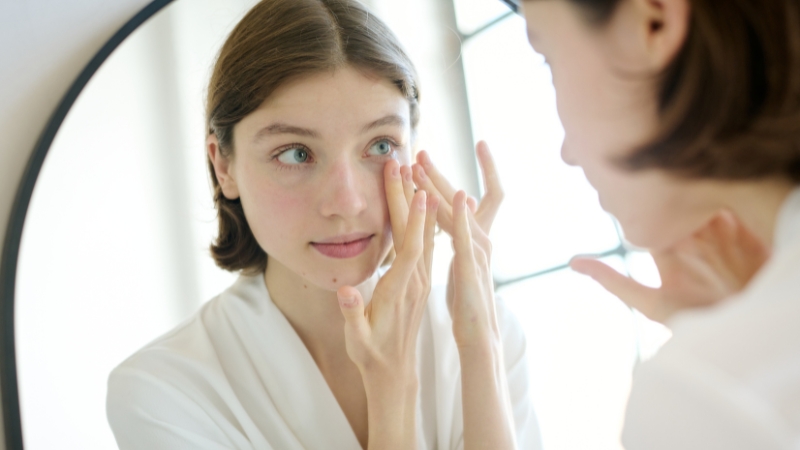 Mlada žena nanosi kremu na područje oko očiju dok se gleda u ogledalo