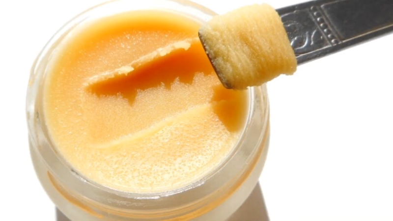 Otvorena teglica Rêve de miel balzama za usne sa metalnom špatulom koja zahvata gustu, medastu teksturu