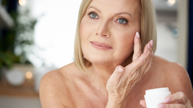 Žena srednjih godina nanosi kremu na lice dok drži otvorenu posudicu sa kremom u ruci
